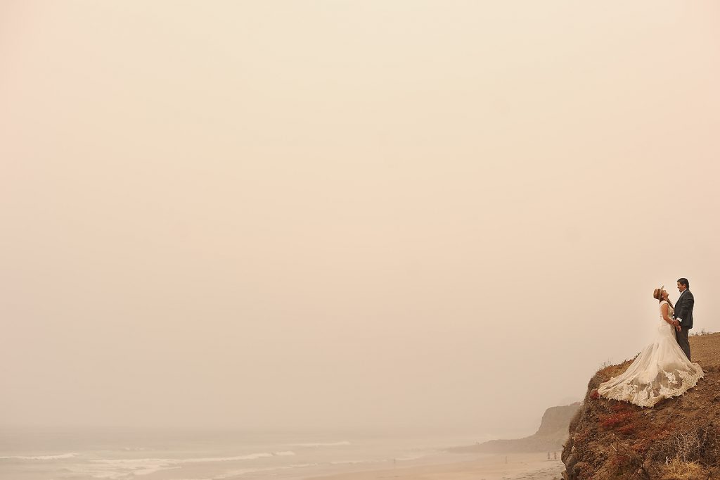 A bride and a groom are standing at the edge of a cliff holding hands and laughing hard during a misty afternoon in an Ensenada beach in Mexico